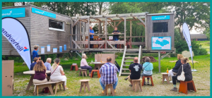 Vor der Forschungsstation, einem mobilen Tiny House, sitzen lokale Bürger:innen und hören der Eröffnungsrede zu.