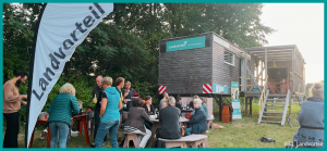 Eine Gruppe von Menschen steht vor einem mobilen Container und unterhält sich. Im Vordergrund steht ein Landvorteil-Banner.