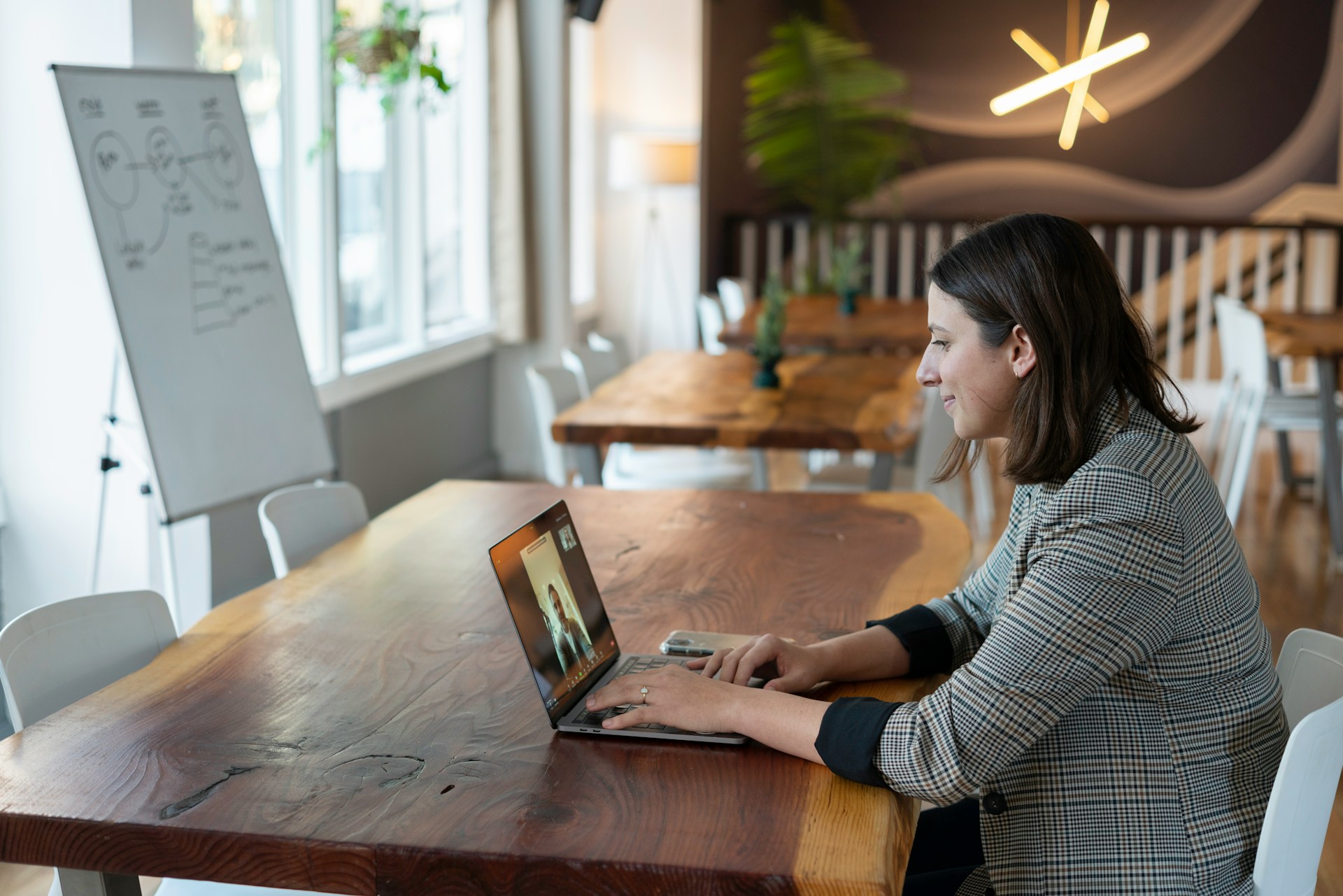 Eine Frau sitzt in einem modernen, hellen Arbeitsraum an einem langen Holztisch und nimmt über ihren Laptop an einer Videokonferenz teil. Im Hintergrund stehen ein Flipchart, weitere Tische und Stühle sowie Pflanzen.