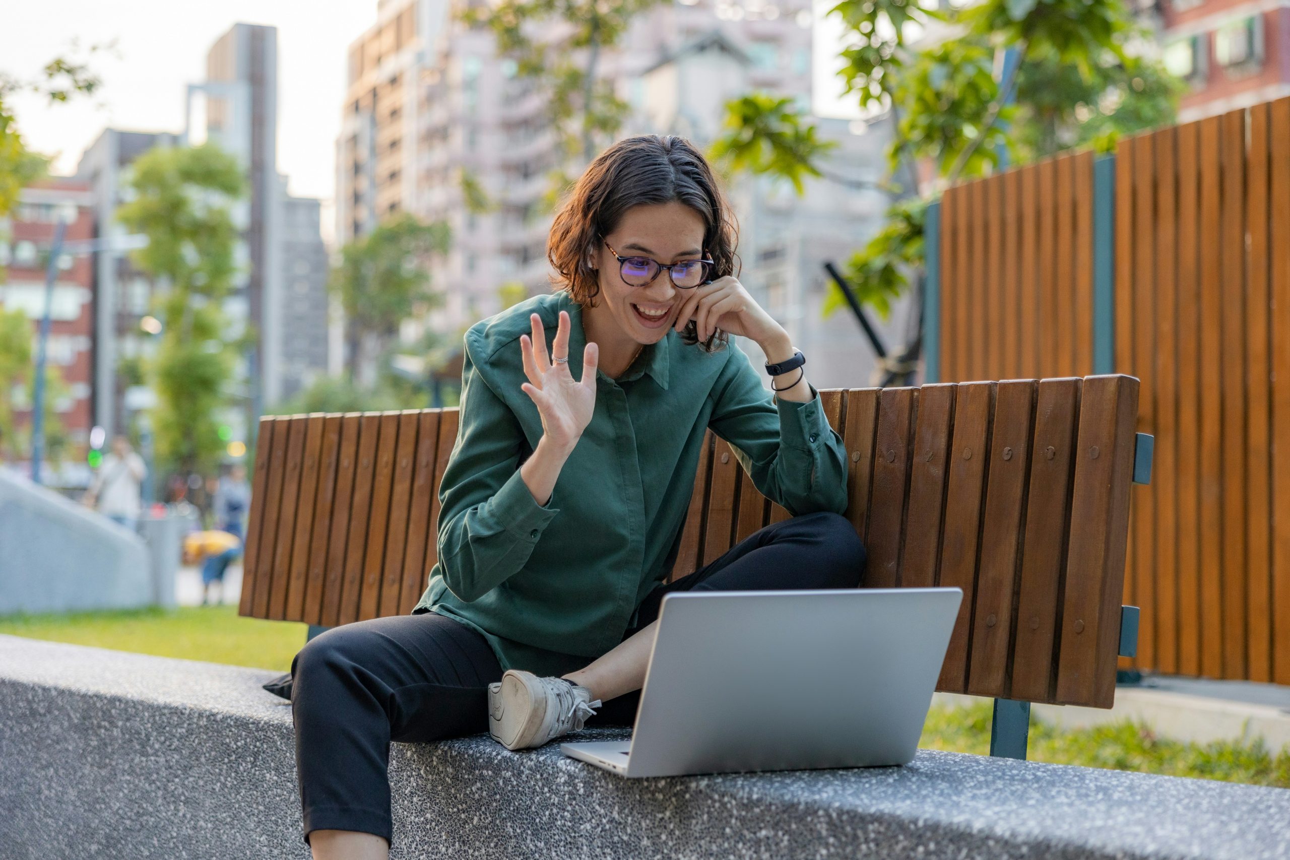 Eine Frau sitzt mit einem Laptop auf einer Holzbank im Freien und winkt lächelnd in die Kamera. Im Hintergrund sind Bäume und moderne Gebäude zu sehen.